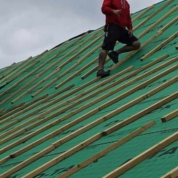 Usługi dekarskie Marcin Jankowski - Dekarz w czerwonej bluzie na zielonym dachu z łatami, widok z dołu pod kątem, pochmurne niebo w tle.