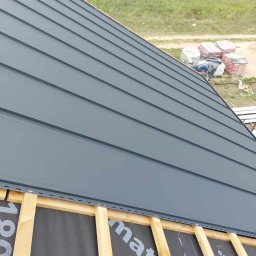 Usługi dekarskie Marcin Jankowski - Montaż szarego dachu panelowego na drewnianej konstrukcji, widoczna membrana dachowa i zapas materiałów budowlanych w tle.