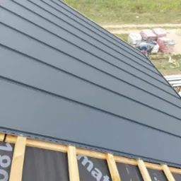 Montaż szarego dachu panelowego na drewnianej konstrukcji, widoczna membrana dachowa i zapas materiałów budowlanych w tle.