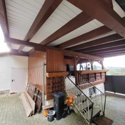 Usługi dekarskie Marcin Jankowski - Zadaszone wejście do budynku z widoczną konstrukcją dachu z drewnianymi belkami i białym poszyciem, schody z metalową balustradą prowadzące na górę, śmietnik i elementy dekoracyjne przy wejściu.