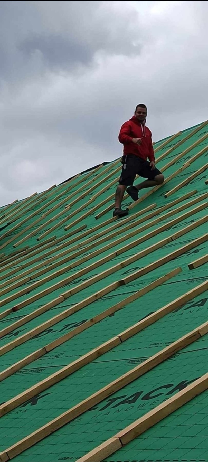 Dekarz w czerwonej bluzie na zielonym dachu z łatami, widok z dołu pod kątem, pochmurne niebo w tle.