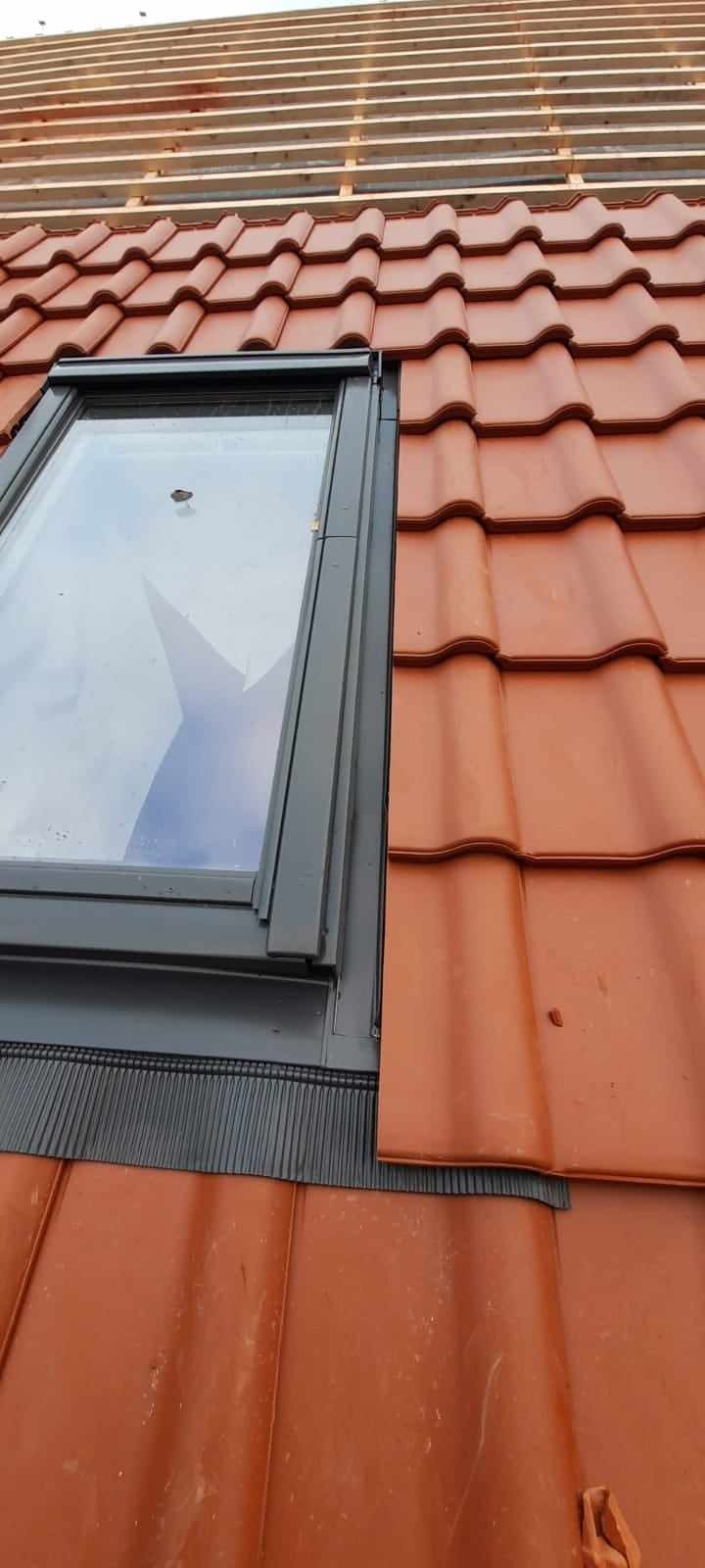 Fragment dachu z dachówki ceramicznej z zamontowanym oknem dachowym z czarną obróbką blacharską i widoczną łatą dachową w tle.