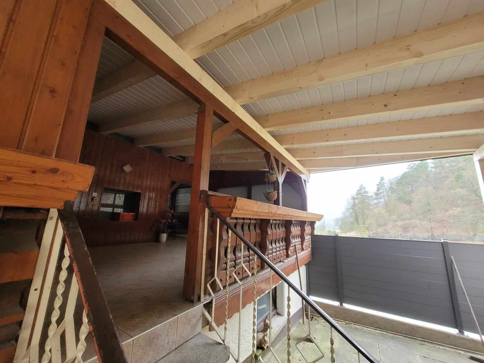 Drewniana konstrukcja dachu z widocznymi belkami i deskami, częściowo zadaszona blachą trapezową, z elementami drewnianej balustrady i schodów, widok z zewnątrz na szary płot i drzewa w tle.