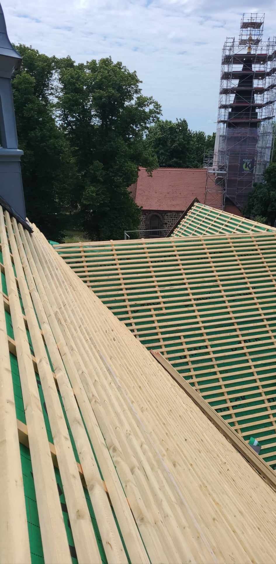 Konstrukcja drewniana dachu z zieloną membraną w tle z widoczną wieżą kościoła w rusztowaniach.