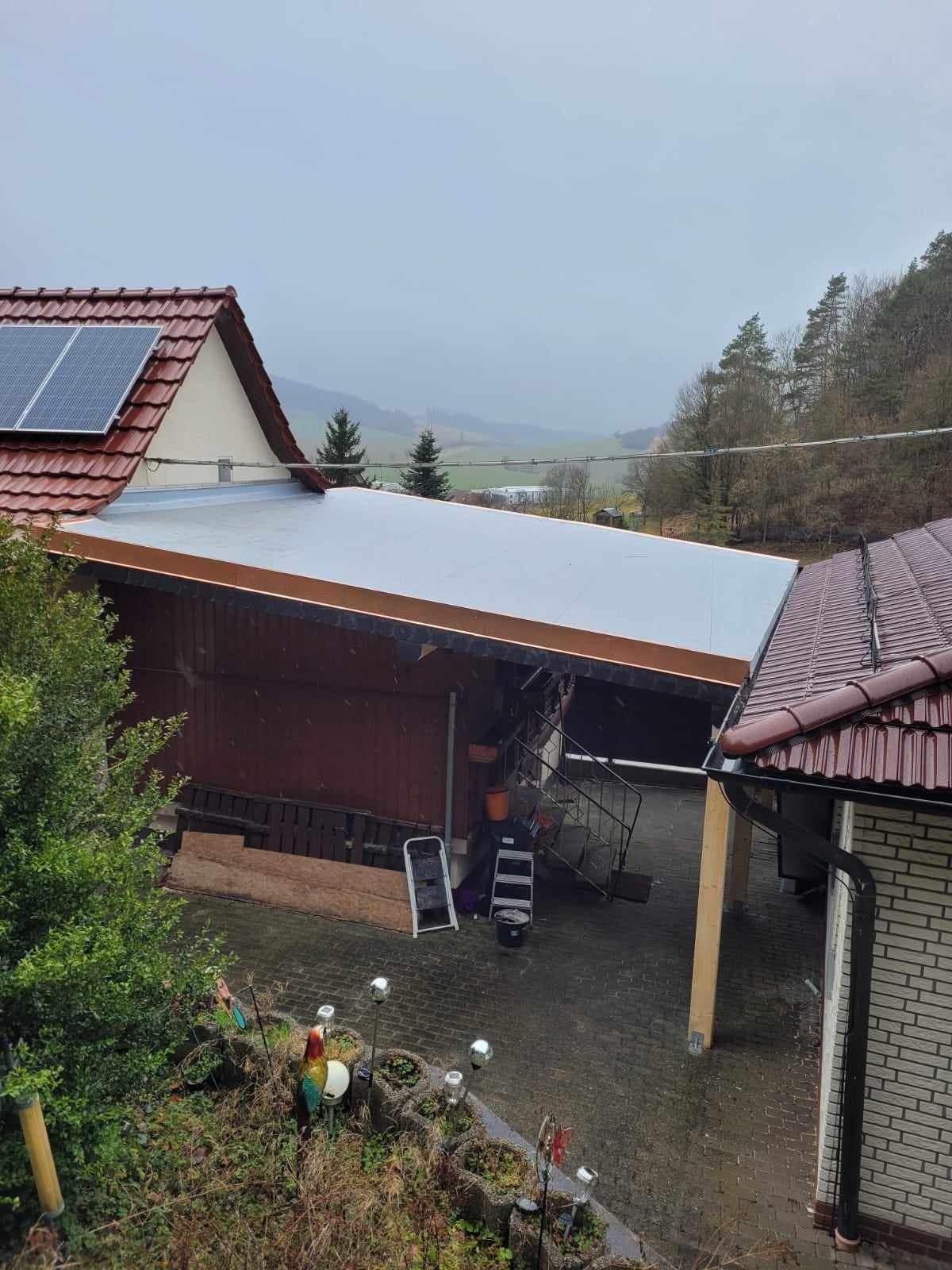 Widok z góry na budynek z płaskim dachem pokrytym jasną membraną, obok dach z panelami słonecznymi i dachówką ceramiczną, w tle zalesione wzgórza w pochmurny dzień.