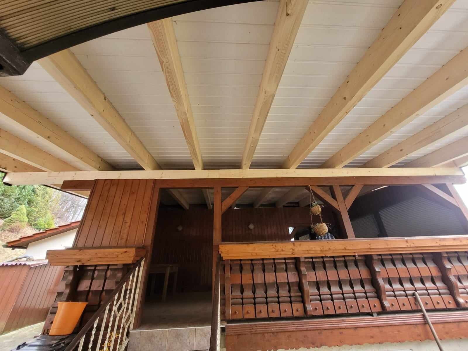 Konstrukcja drewniana nowego zadaszenia tarasu z widocznymi belkami i białą podsufitką, widok z dołu na drewniane balustrady.