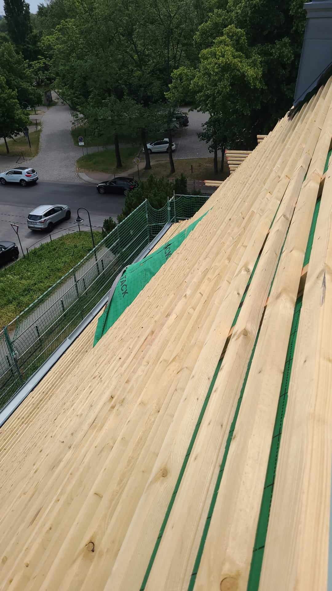 Konstrukcja drewniana dachu w trakcie budowy, widoczne deski, membrana dachowa i siatka zabezpieczająca, w tle ulica z samochodami i drzewami.