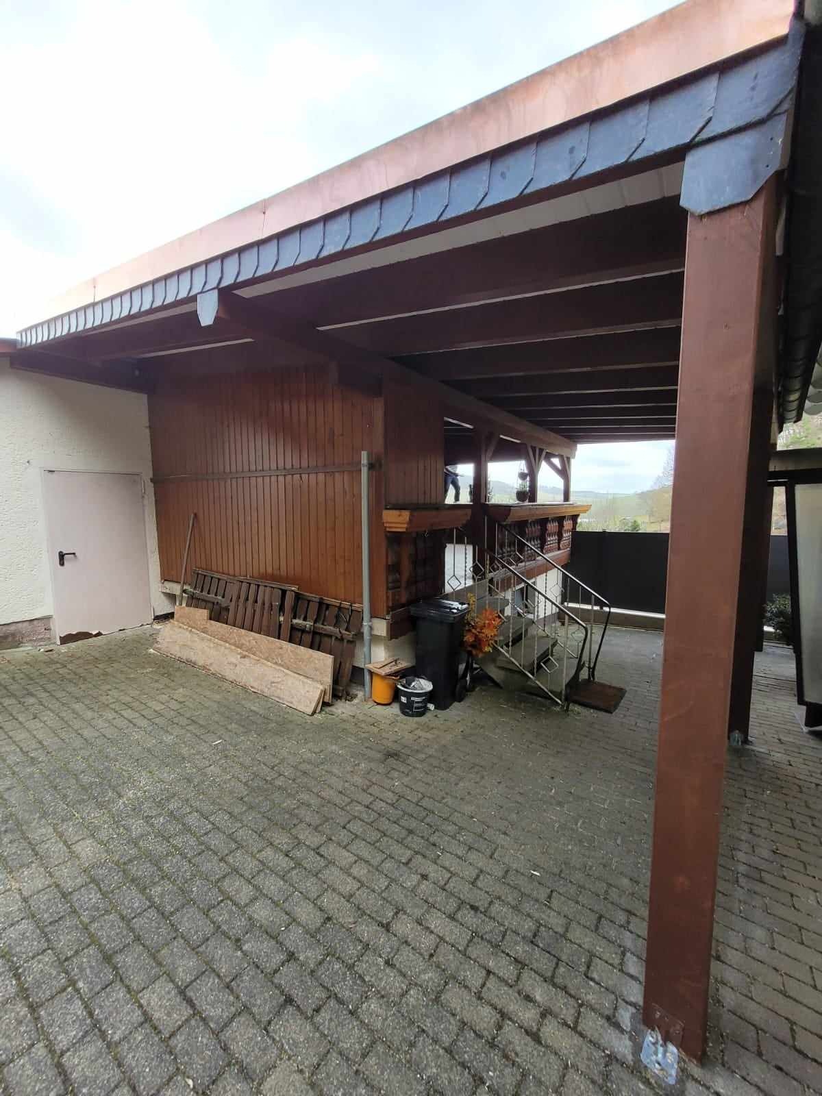 Zadaszenie z drewnianą konstrukcją i fragmentem dachu pokrytego szarymi płytkami, widoczne także schody z metalową barierką i elementy otoczenia.