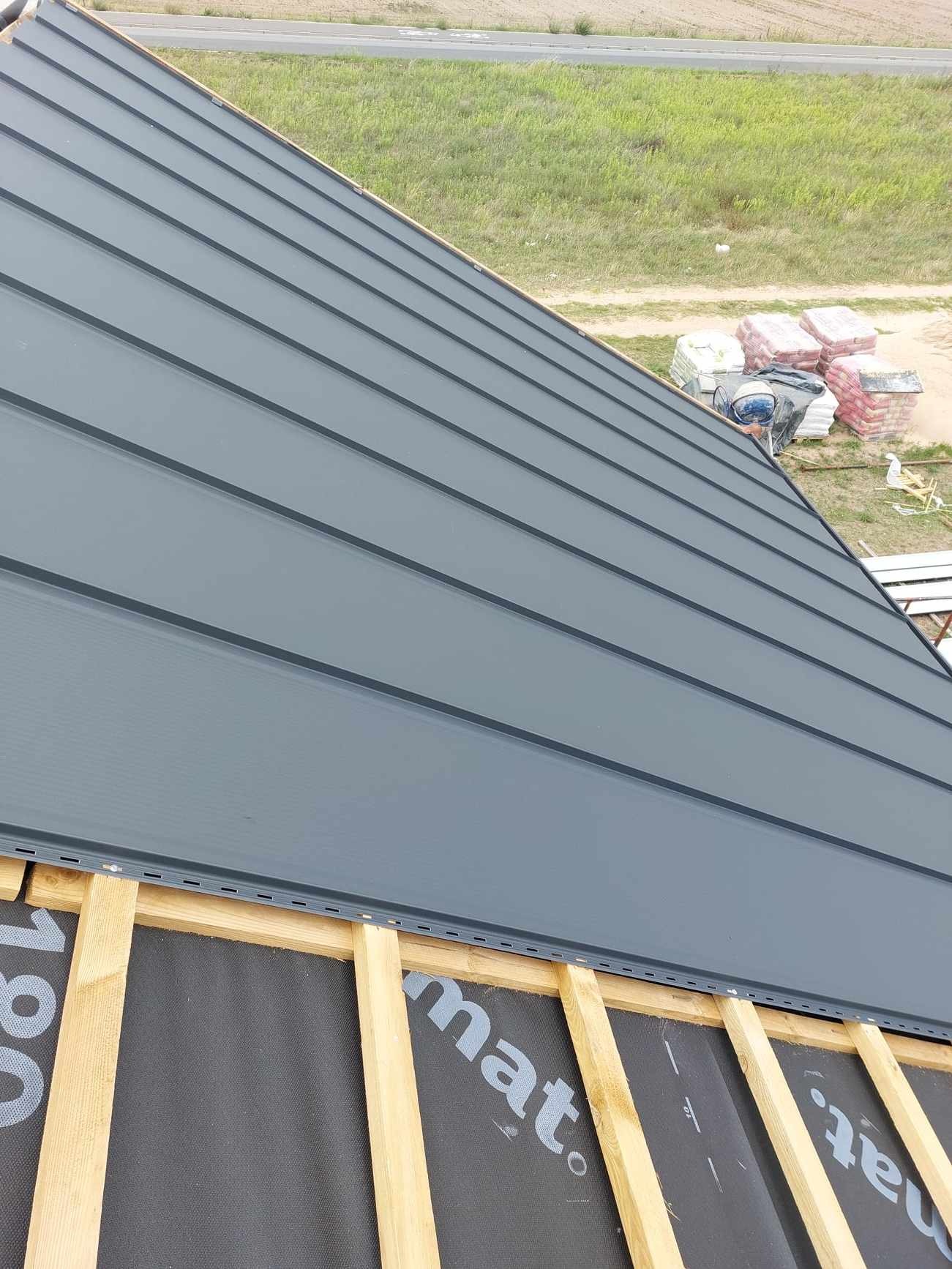 Montaż szarego dachu panelowego na drewnianej konstrukcji, widoczna membrana dachowa i zapas materiałów budowlanych w tle.