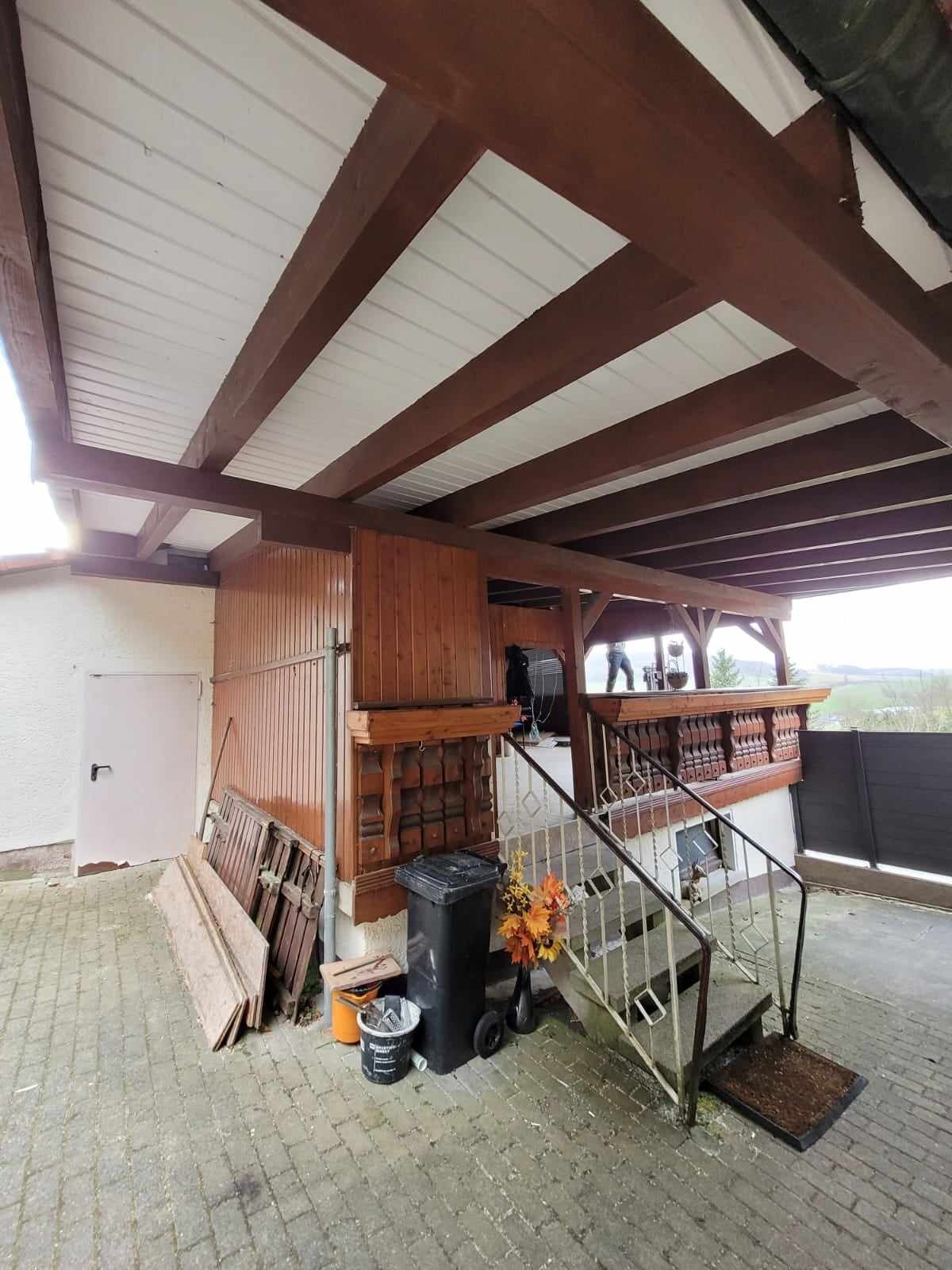 Zadaszone wejście do budynku z widoczną konstrukcją dachu z drewnianymi belkami i białym poszyciem, schody z metalową balustradą prowadzące na górę, śmietnik i elementy dekoracyjne przy wejściu.