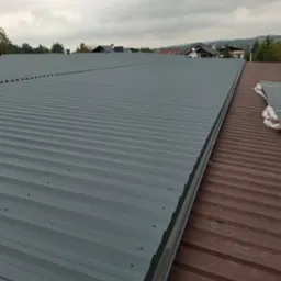 Nowy, szary dach z blachy trapezowej, kontrastujący z fragmentem starego, brązowego dachu, widok z góry na tle pochmurnego nieba i zabudowań.