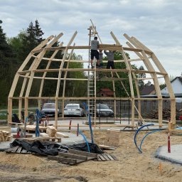 Tomasz Mikołajczyk TM Dachy-Daszki - Budowa drewnianej konstrukcji kopuły przez trzech mężczyzn na tle drzew i domów. Widoczne samochody i materiały budowlane.