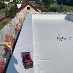 Tomasz Mikołajczyk TM Dachy-Daszki - Biała, płaska powierzchnia dachu pokryta membraną, z widocznym czerwonym laserem pomiarowym. W tle budynki mieszkalne i zieleń. Widok z góry, w słoneczny dzień.