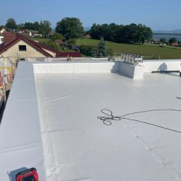 Tomasz Mikołajczyk TM Dachy-Daszki - Biała, płaska powierzchnia dachu po hydroizolacji z widocznymi kominami i pracownikiem z dmuchawą na tle krajobrazu z jeziorem i zabudowaniami.