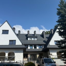 Tomasz Mikołajczyk TM Dachy-Daszki - Biały budynek z ciemnoszarym dachem z blachy na tle błękitnego nieba. Widoczne rusztowania i samochody, sugerujące prace wykończeniowe.