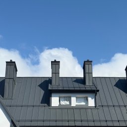 Tomasz Mikołajczyk TM Dachy-Daszki - Szary dach z blachy na białym budynku z trzema kominami na tle błękitnego nieba z chmurami. Okna na poddaszu zabezpieczone folią.