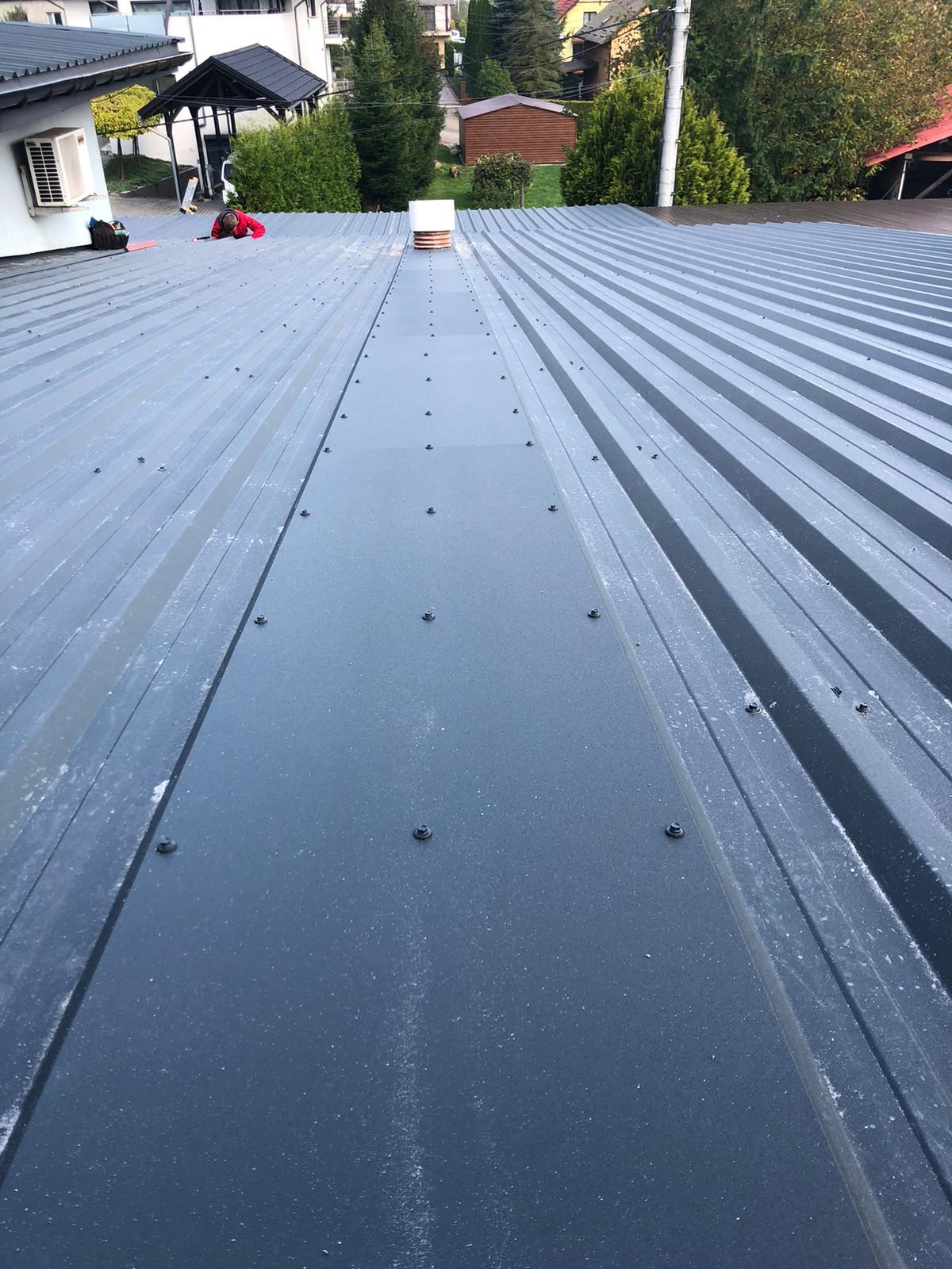 Szary dach z blachy trapezowej, widoczne łączenia paneli i śruby mocujące, w tle budynki mieszkalne i pracownik w czerwonej kurtce.