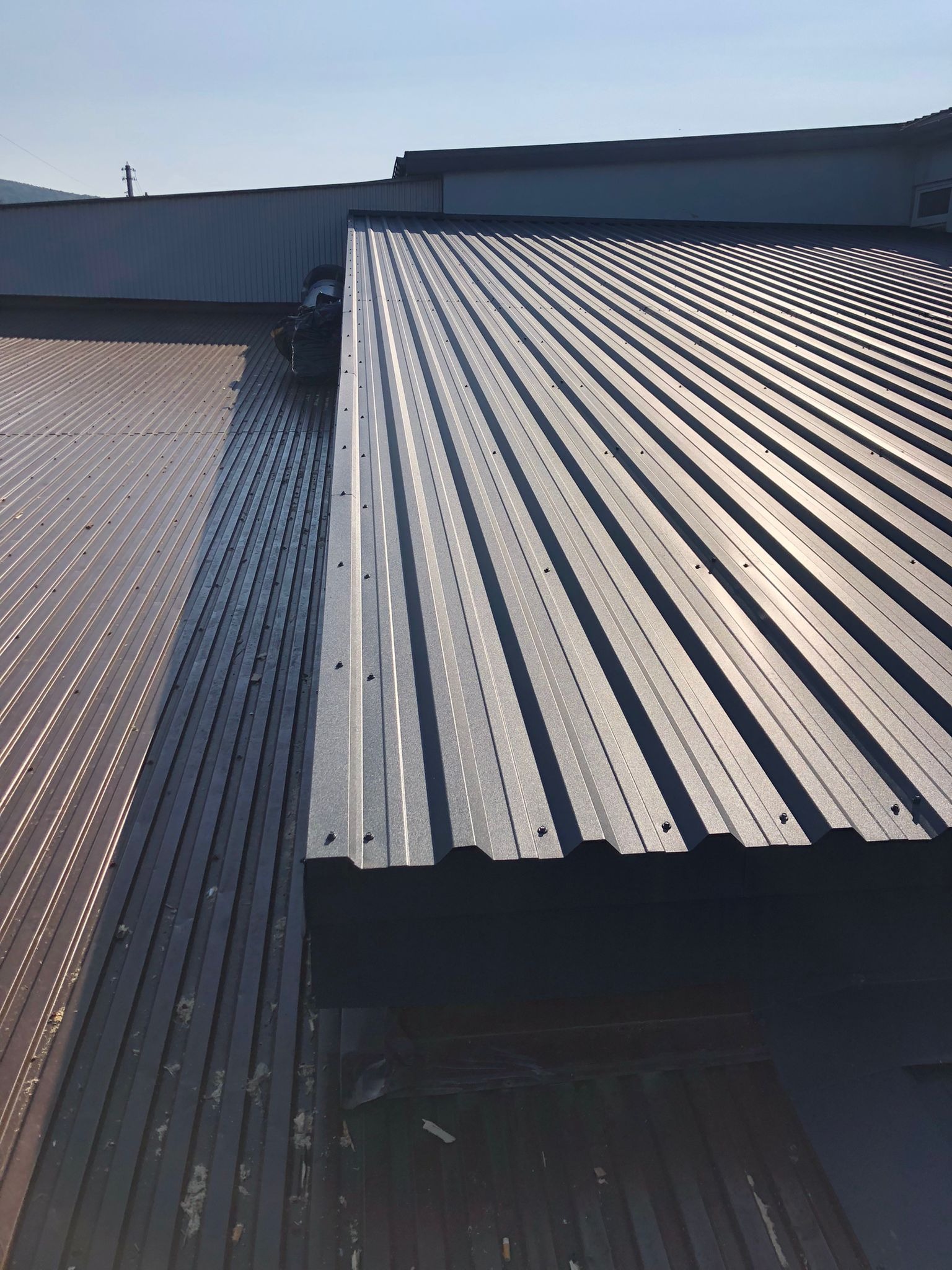 Fragment dachu z blachy trapezowej w trakcie wymiany; widoczna stara, zniszczona część dachu obok nowej, błyszczącej blachy z widocznymi śrubami mocującymi.
