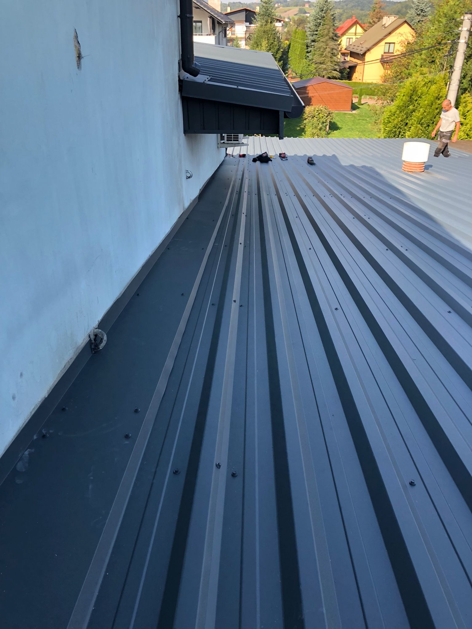 Nowy, szary dach panelowy z widocznym systemem mocowań, kominkiem wentylacyjnym i dekarzem w oddali, na tle zabudowy mieszkalnej.