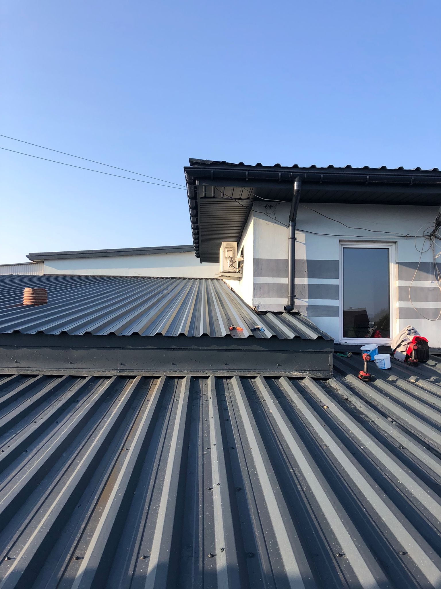 Szary dach z blachy trapezowej, widoczne narzędzia pracy, wiadro z farbą i plecak na dachu, fragment budynku z oknem i rynną w tle.