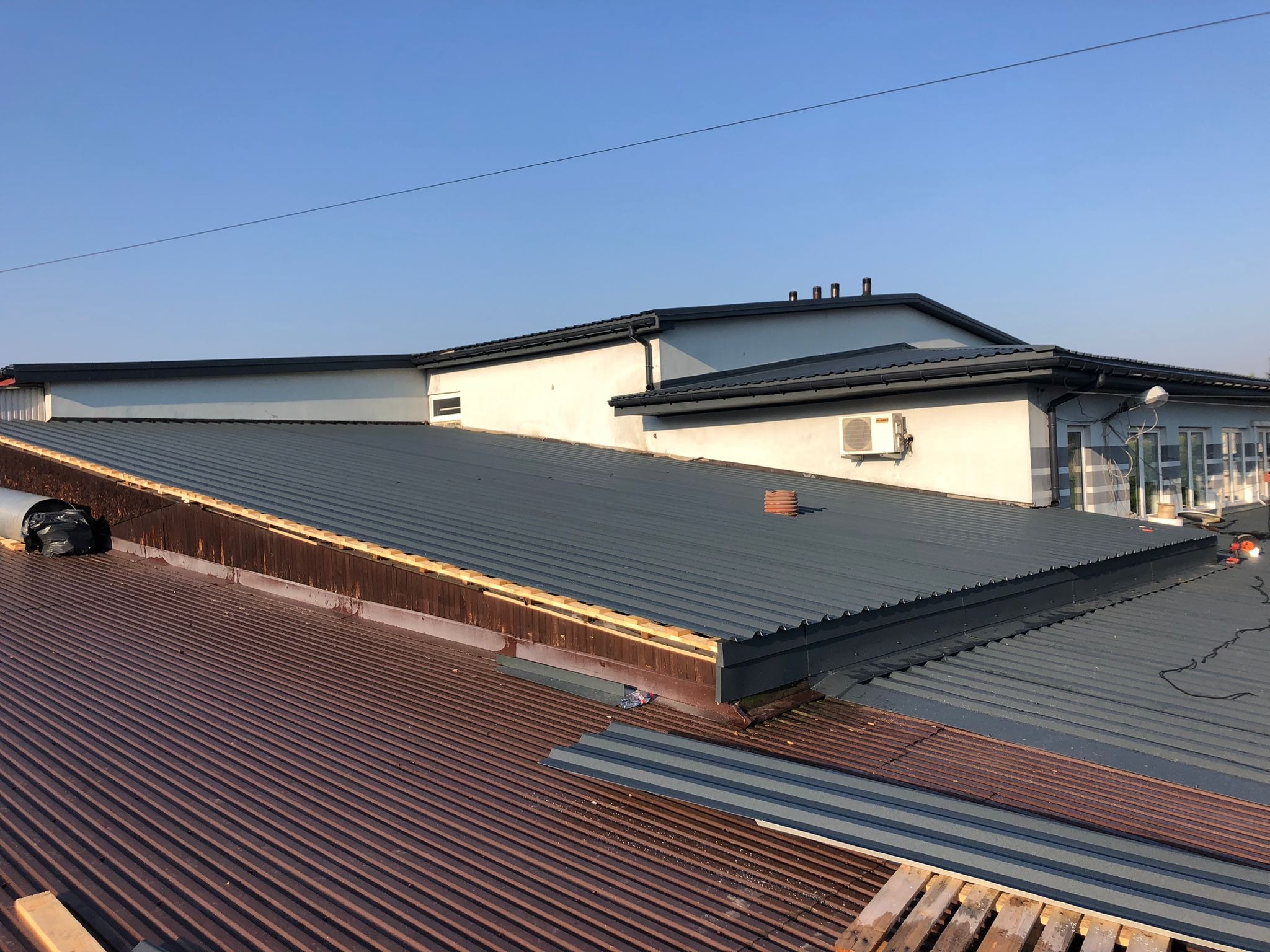 Fragment dachu budynku z blachy trapezowej w kolorze brązowym i szarym, widoczne elementy konstrukcji drewnianej, w tle budynek z klimatyzatorem na ścianie, błękitne niebo.