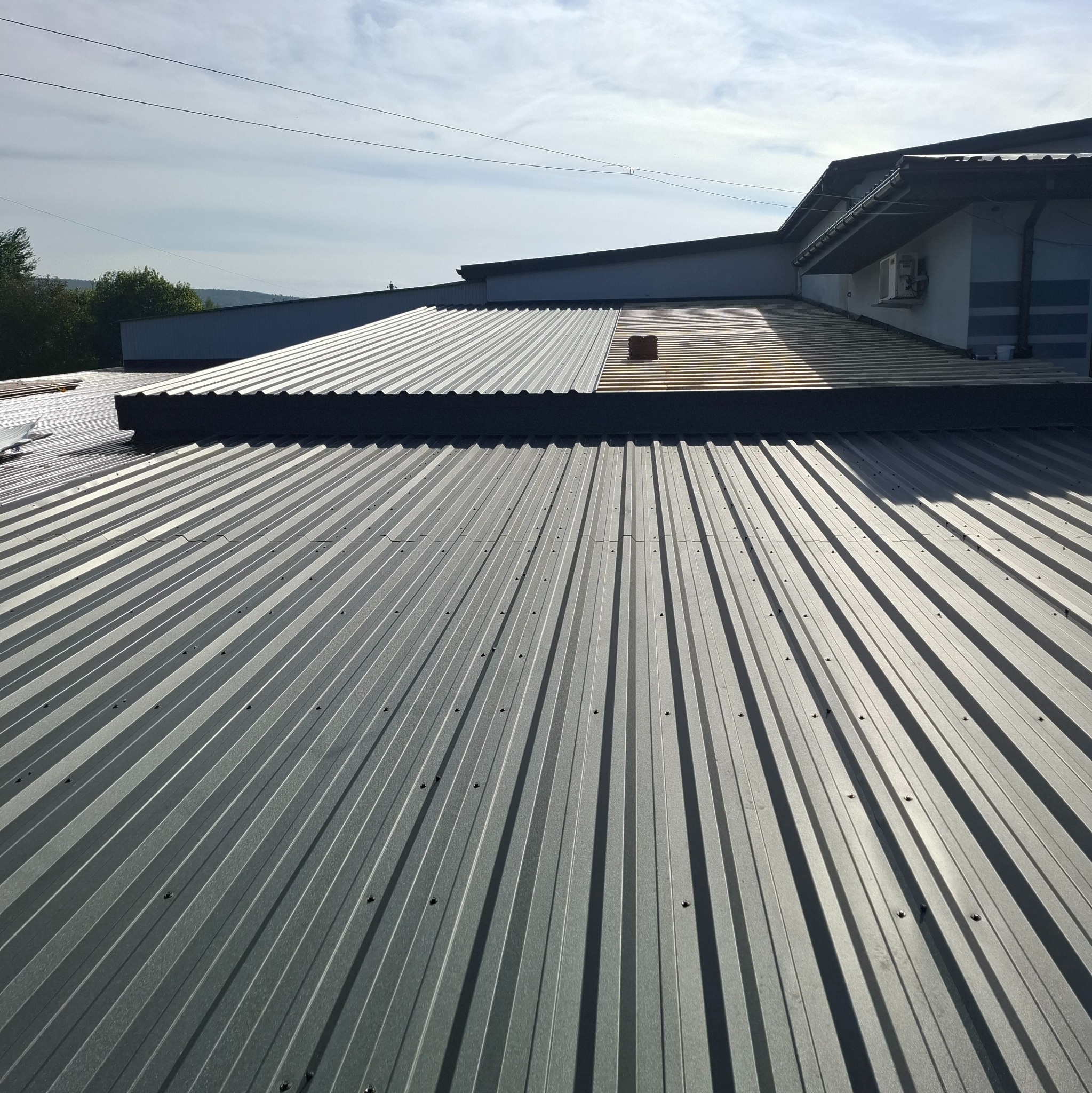 Nowy, blaszany dach trapezowy w kolorze grafitowym z widocznymi mocowaniami, częściowo w cieniu, z fragmentem starego dachu i kominem w tle.