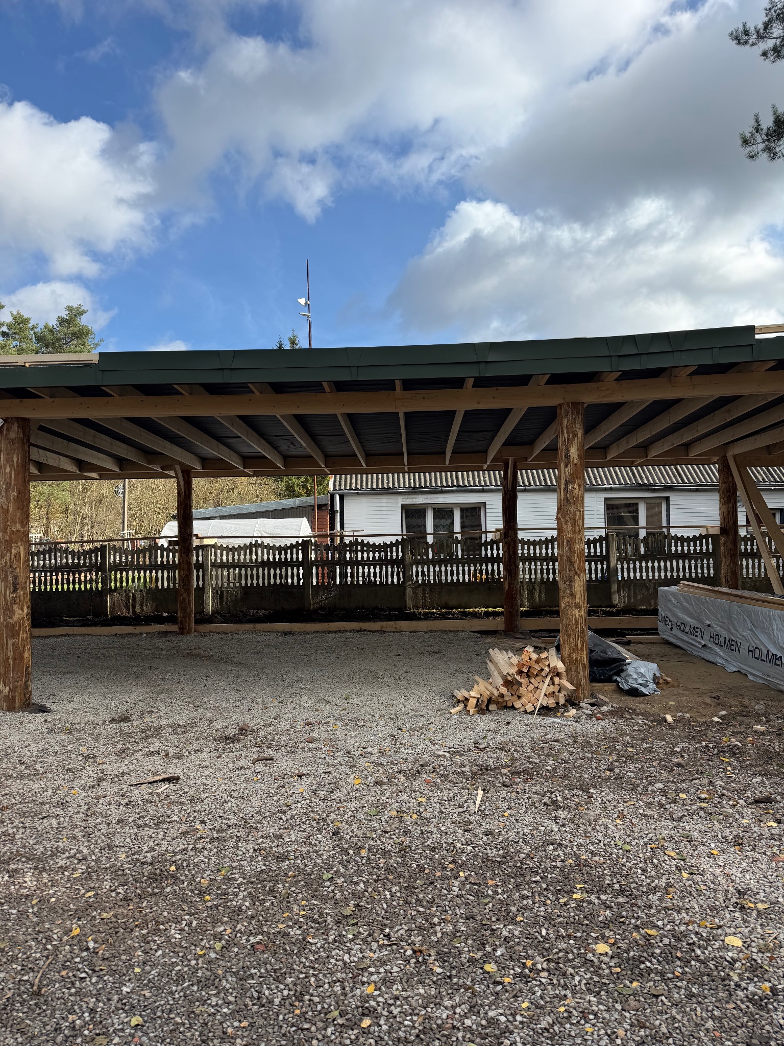 Konstrukcja z drewnianych bali pod zadaszenie, widoczne materiały budowlane i niebo z chmurami. Budowa w toku, surowe drewno, teren przygotowany pod dalsze prace.