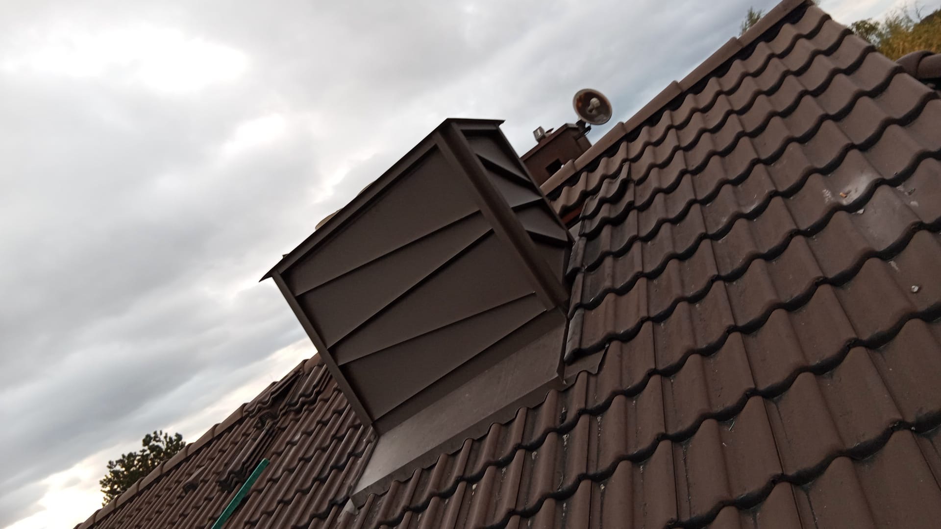 Brązowy dach z dachówki ceramicznej z nowoczesnym, metalowym wyłazem dachowym na tle pochmurnego nieba. Ujęcie z dołu, podkreślające detale.