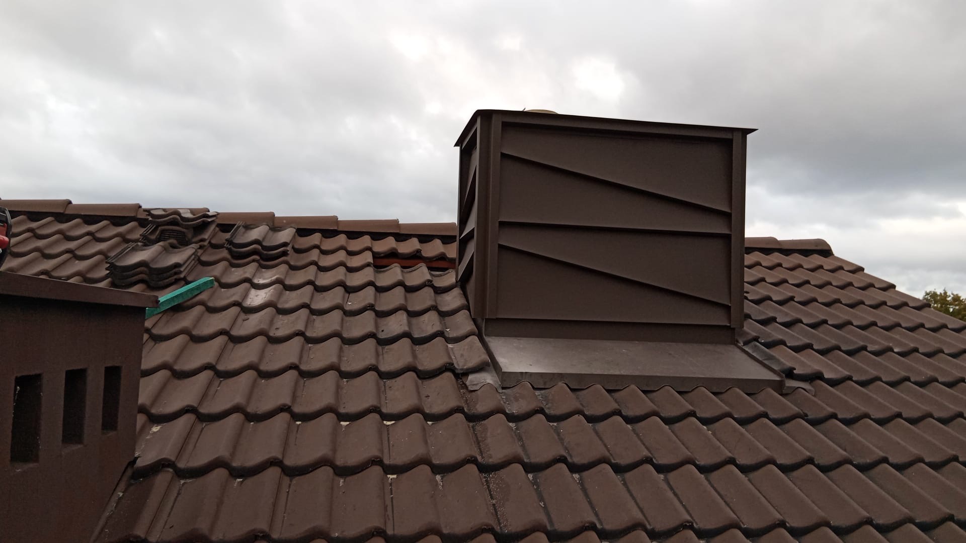 Brązowy dach z dachówki ceramicznej z nową obróbką komina z blachy w kolorze dachu. Część dachówek zdjęta, przygotowane do wymiany. Pochmurne niebo w tle.