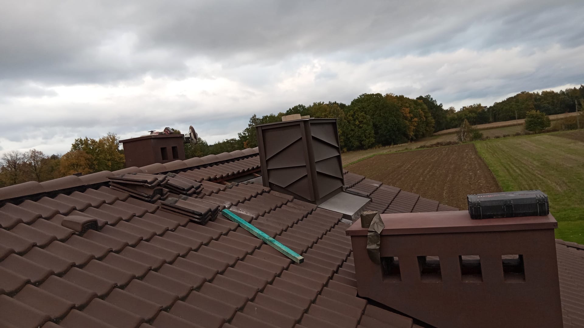 Brązowy dach z dachówki ceramicznej, komin z obróbką blacharską, łatą i narzędziami na tle pochmurnego nieba i pól uprawnych. Widok z góry.