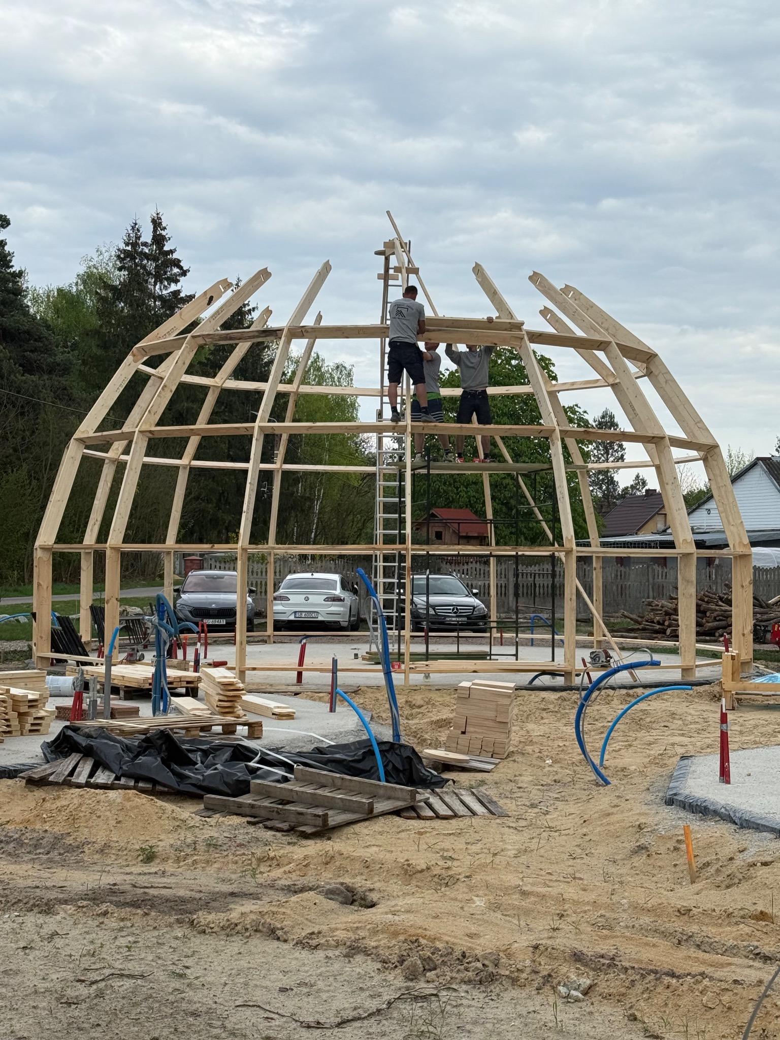 Budowa drewnianej konstrukcji kopuły przez trzech mężczyzn na tle drzew i domów. Widoczne samochody i materiały budowlane.