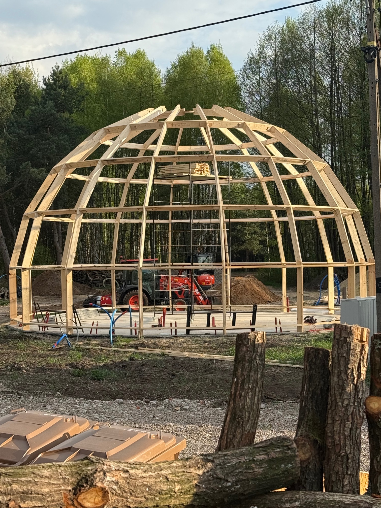 Konstrukcja drewniana kopuły w trakcie budowy, widoczny szkielet z belek na betonowej podstawie, w tle las i sprzęt budowlany. Ujęcie z dołu, widać fragmenty pni drzew.