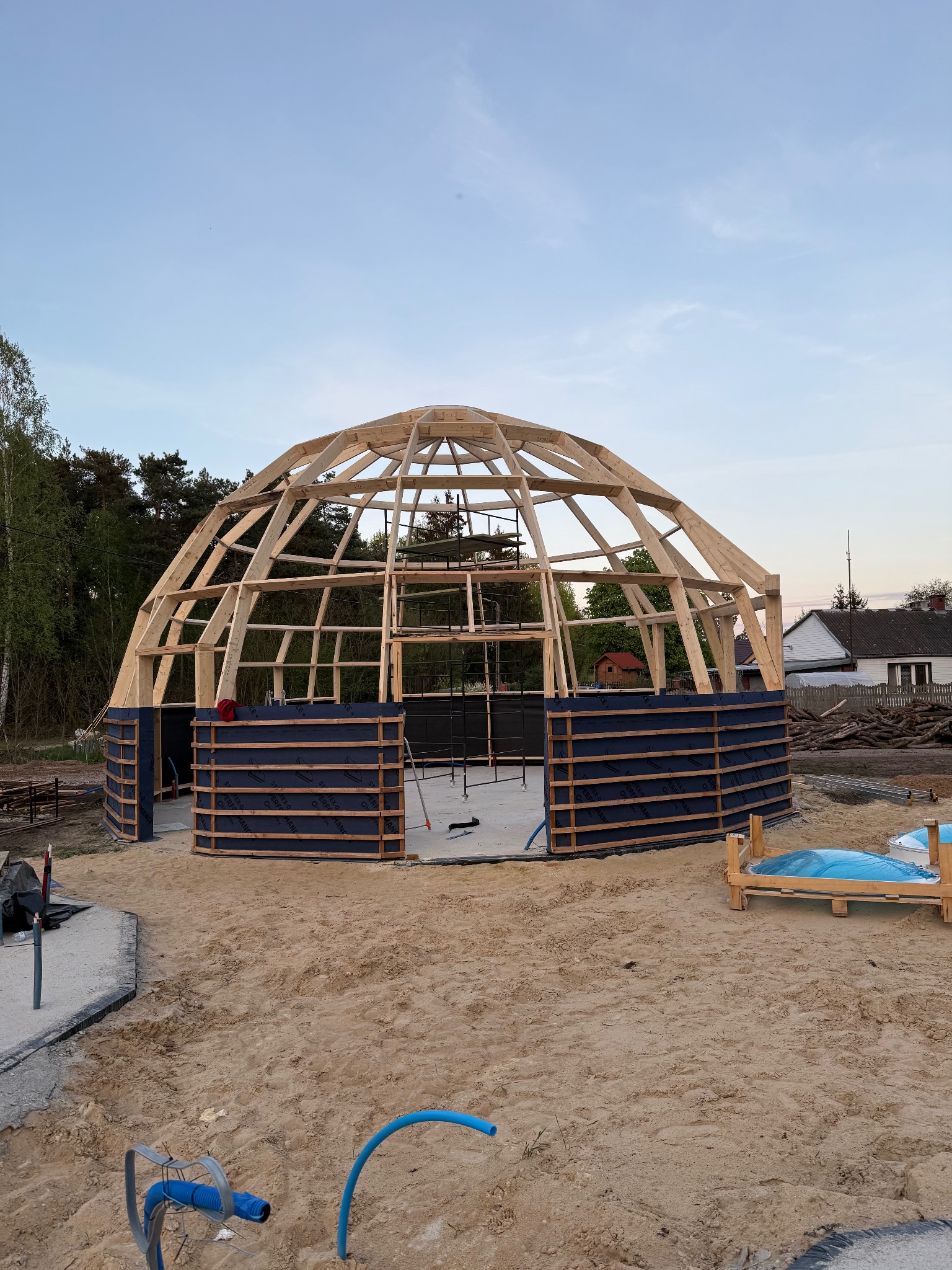 Konstrukcja drewniana kopuły w trakcie budowy, widoczne elementy więźby i izolacji, teren budowy z piaskiem i fragmentami materiałów budowlanych. Ujęcie z dołu.
