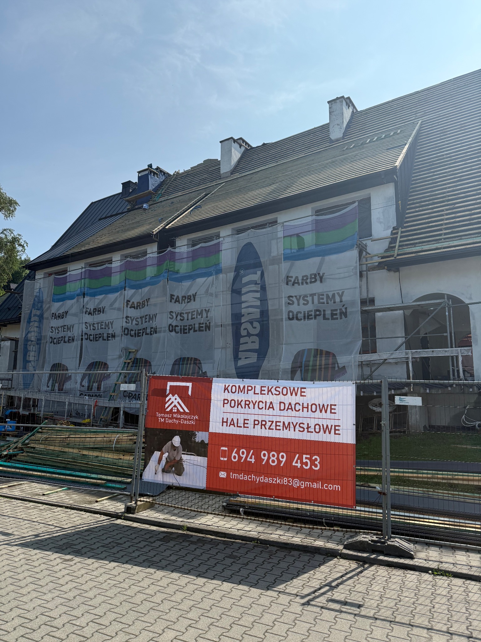 Budynek w trakcie remontu dachu, zabezpieczony siatką z reklamą farb i systemów ociepleń. Na pierwszym planie baner firmy dekarskiej z numerem telefonu.