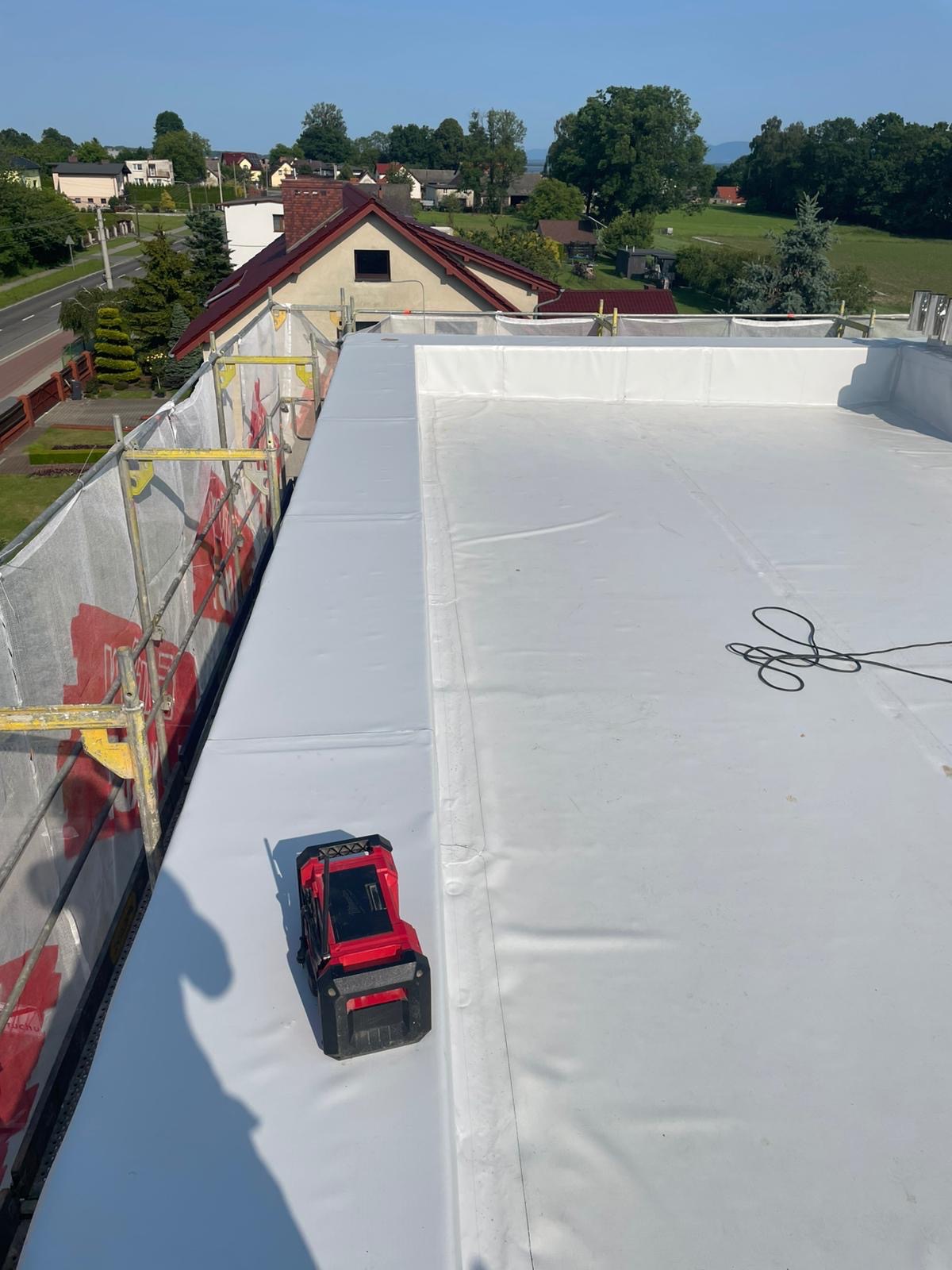 Biała, płaska powierzchnia dachu pokryta membraną, z widocznym czerwonym laserem pomiarowym. W tle budynki mieszkalne i zieleń. Widok z góry, w słoneczny dzień.