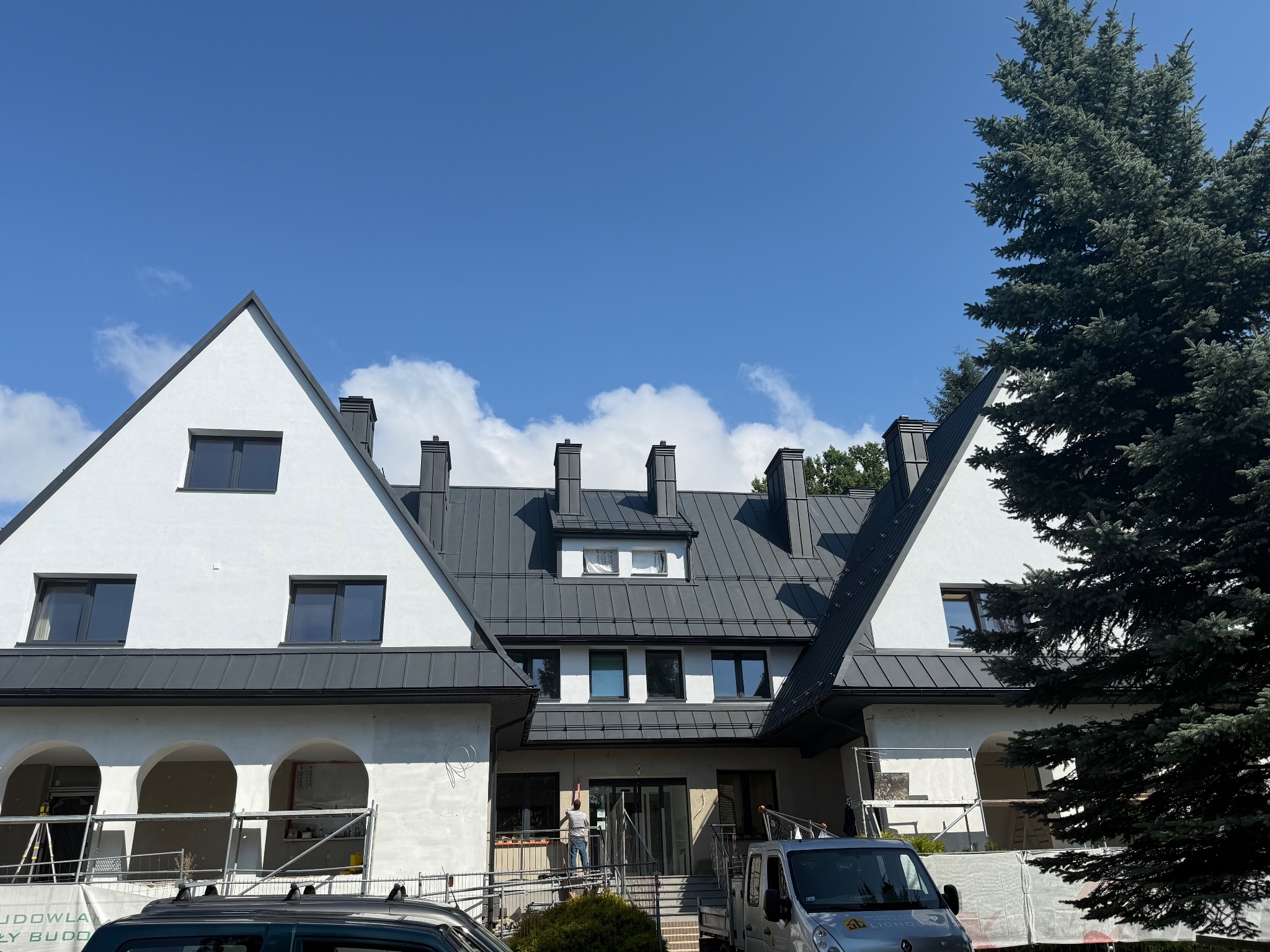 Biały budynek z ciemnoszarym dachem z blachy na tle błękitnego nieba. Widoczne rusztowania i samochody, sugerujące prace wykończeniowe.