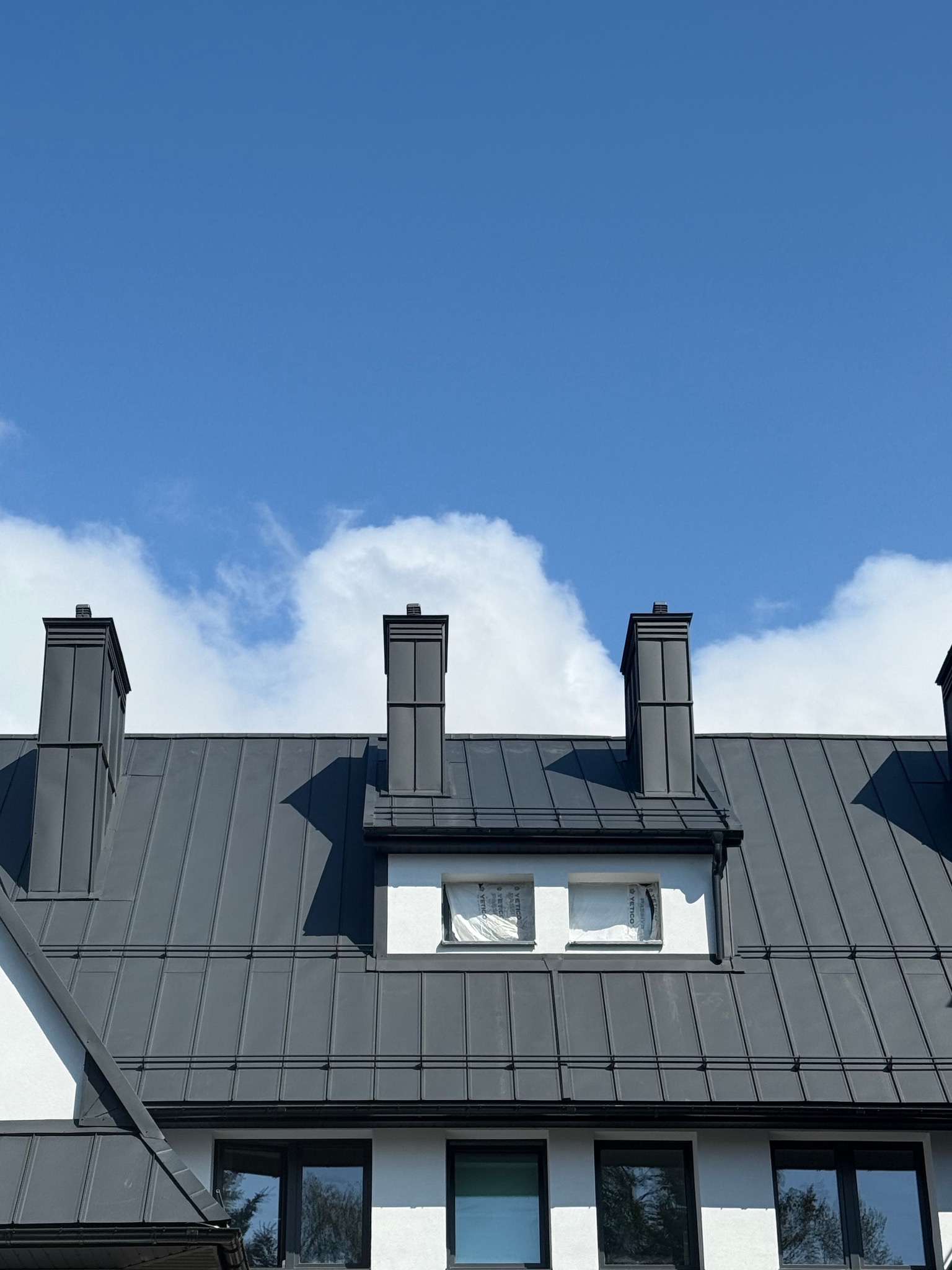 Szary dach z blachy na białym budynku z trzema kominami na tle błękitnego nieba z chmurami. Okna na poddaszu zabezpieczone folią.