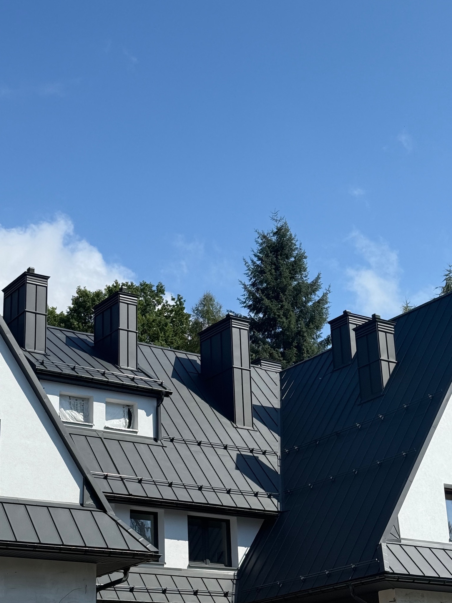 Czarny, metalowy dach z kominami na tle błękitnego nieba i zieleni drzew w Wadowicach. Nowoczesne pokrycie dachowe z blachy na budynku.