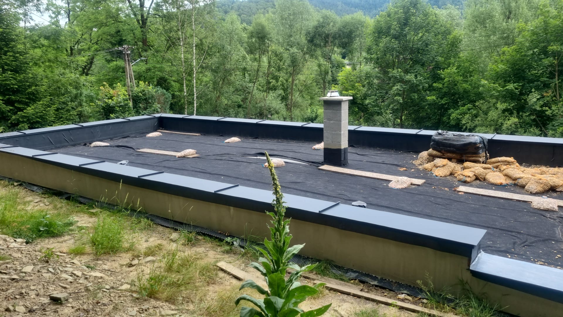 Płaski dach pokryty czarną membraną, z widocznym kominem i workami z materiałem izolacyjnym. W tle zieleń drzew i pagórki. Widok z dołu, z bliska.