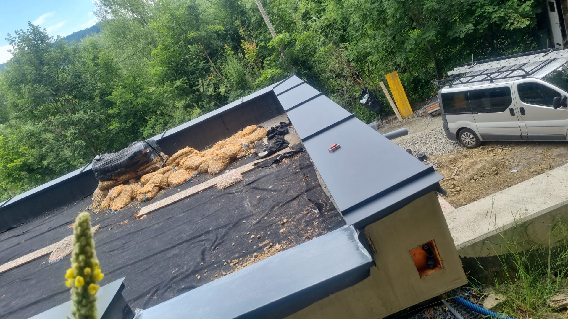 Szary dach z blachy na budynku w trakcie budowy, widoczne worki z materiałem izolacyjnym, w tle zieleń drzew i zaparkowany bus.