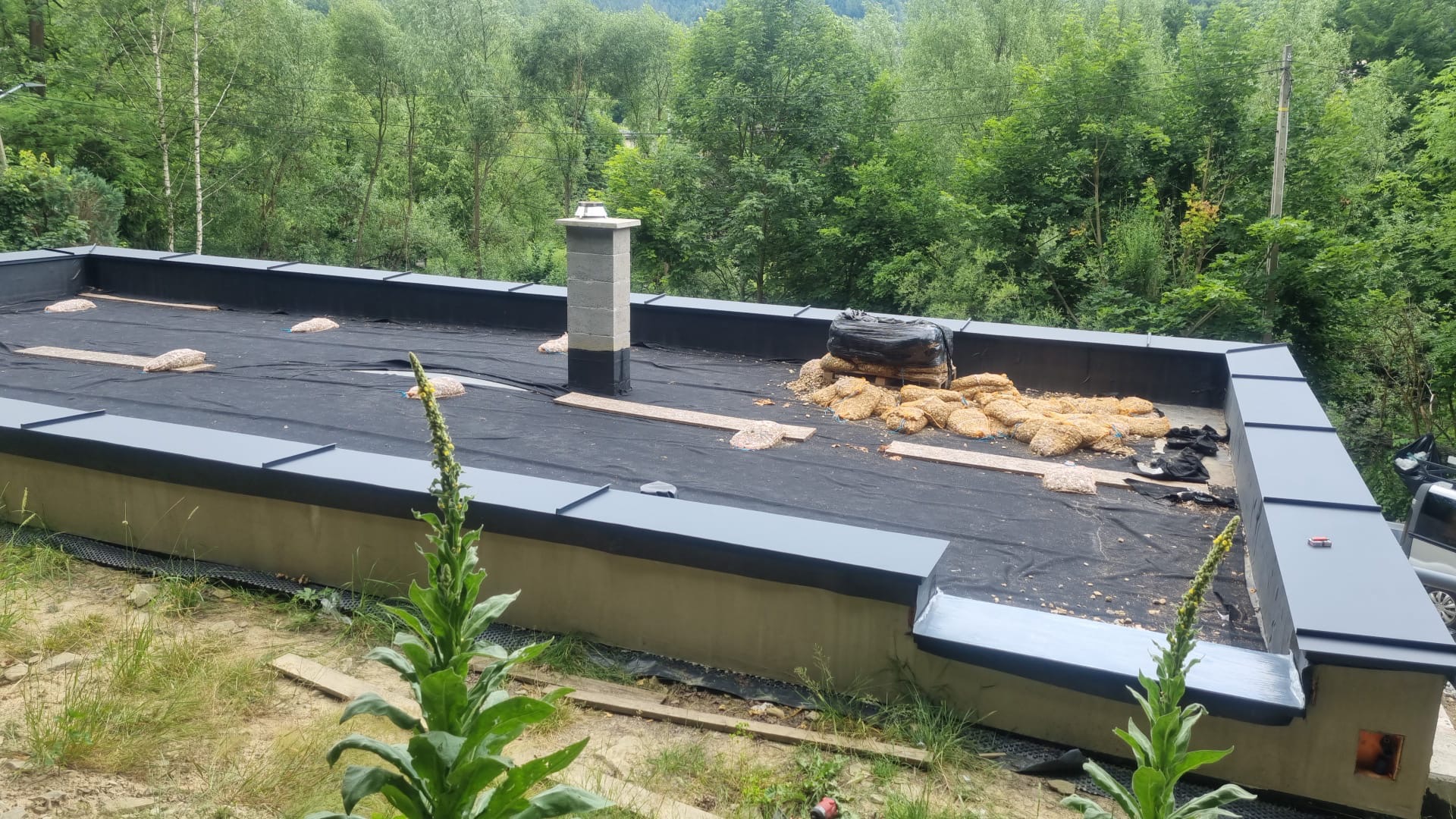 Płaski dach z czarną membraną, obróbkami blacharskimi w kolorze grafitowym i kominem z szarego pustaka. Widoczne worki z materiałem izolacyjnym.