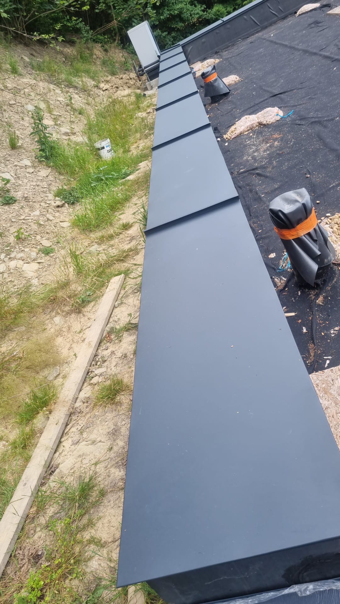 Fragment dachu z nową, grafitową obróbką blacharską, widoczne zabezpieczenia na membranie dachowej i fragment terenu z trawą. Ujęcie z góry, z bliska.