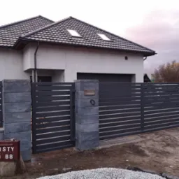 Nowoczesne ogrodzenie panelowe w kolorze grafitowym z przesuwną bramą i furtką, zamontowane przed domem z szarą dachówką, widoczna skrzynka na listy w stylu retro z napisem 'LISTY 5 BB'.