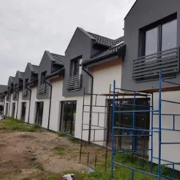 Szereg nowo wybudowanych domów w odcieniach szarości i bieli, z widocznymi rusztowaniami przed jednym z budynków, sugerujące prace wykończeniowe.