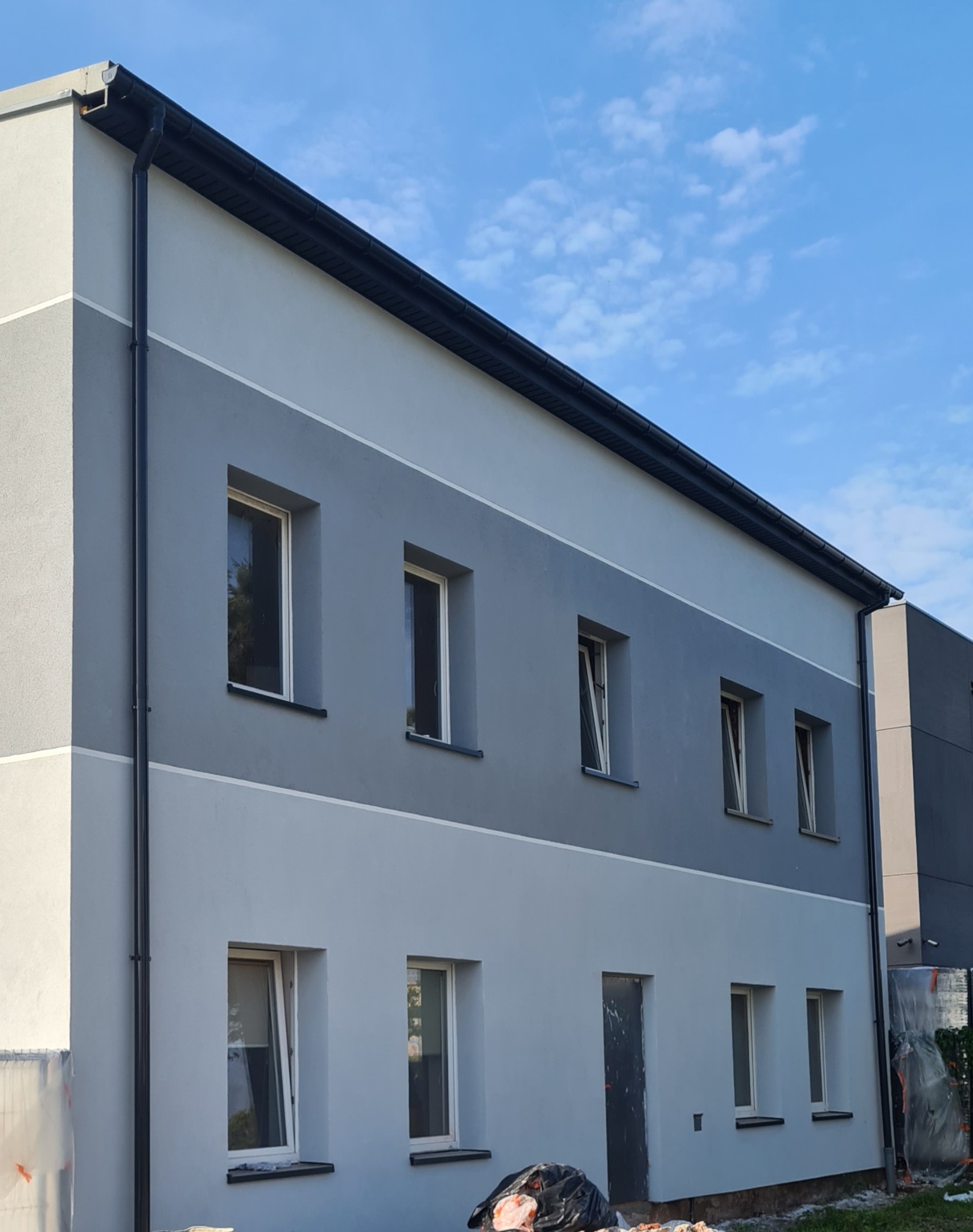 Dwukondygnacyjny budynek po odnowieniu elewacji w odcieniach szarości z widocznymi oknami i czarną rynną, z elementami zabezpieczeń budowlanych na pierwszym planie.