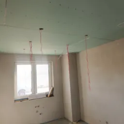 Pokój w trakcie remontu: zamontowana płyta karton gips na suficie z wystającymi przewodami, widoczne ślady po instalacjach na ścianach, okno z widokiem na zewnątrz, na podłodze resztki materiałów...