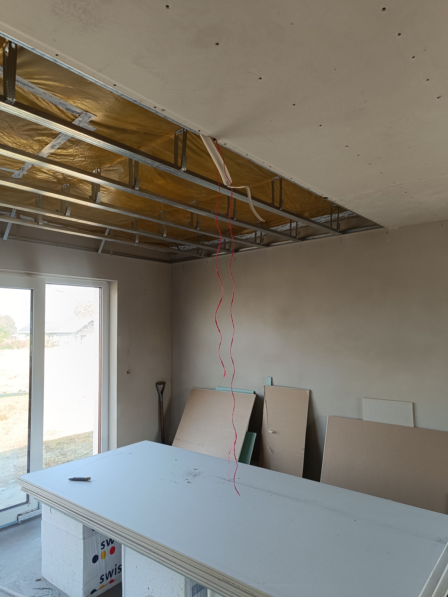 Wnętrze w trakcie remontu: metalowa konstrukcja sufitu podwieszanego z izolacją, przygotowane płyty kartonowo-gipsowe, wiszące kable elektryczne, narzędzie na stosie płyt.