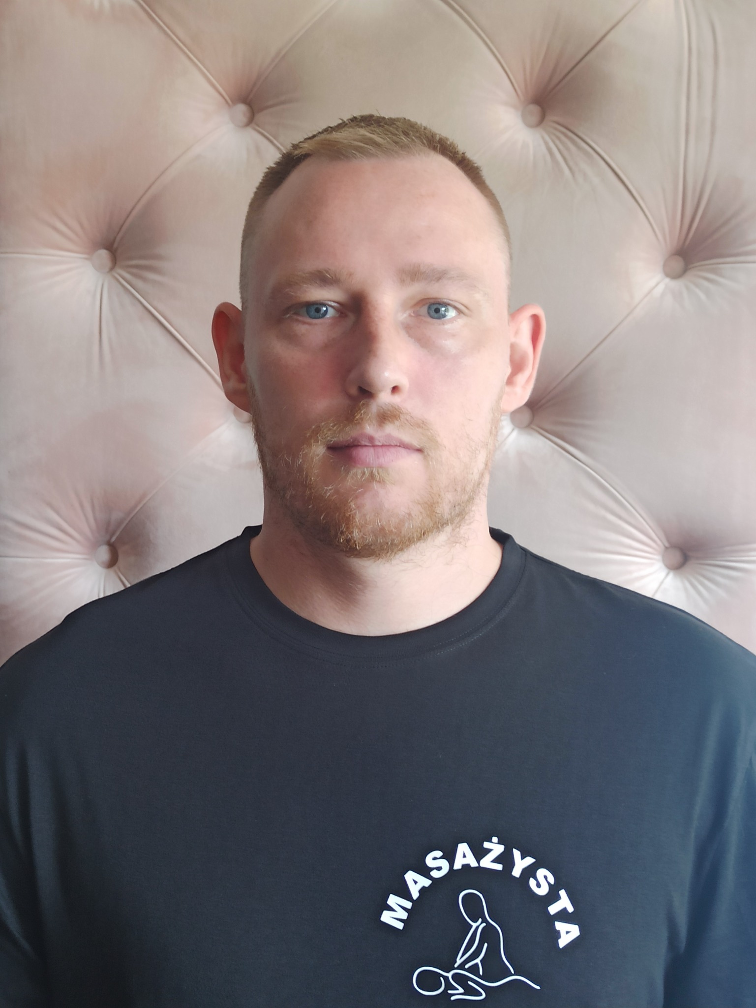 Portret mężczyzny w czarnej koszulce z napisem 'MASAŻYSTA' i prostym logo przedstawiającym osobę podczas zabiegu, na tle tapicerowanej ściany w odcieniach pudrowego różu.