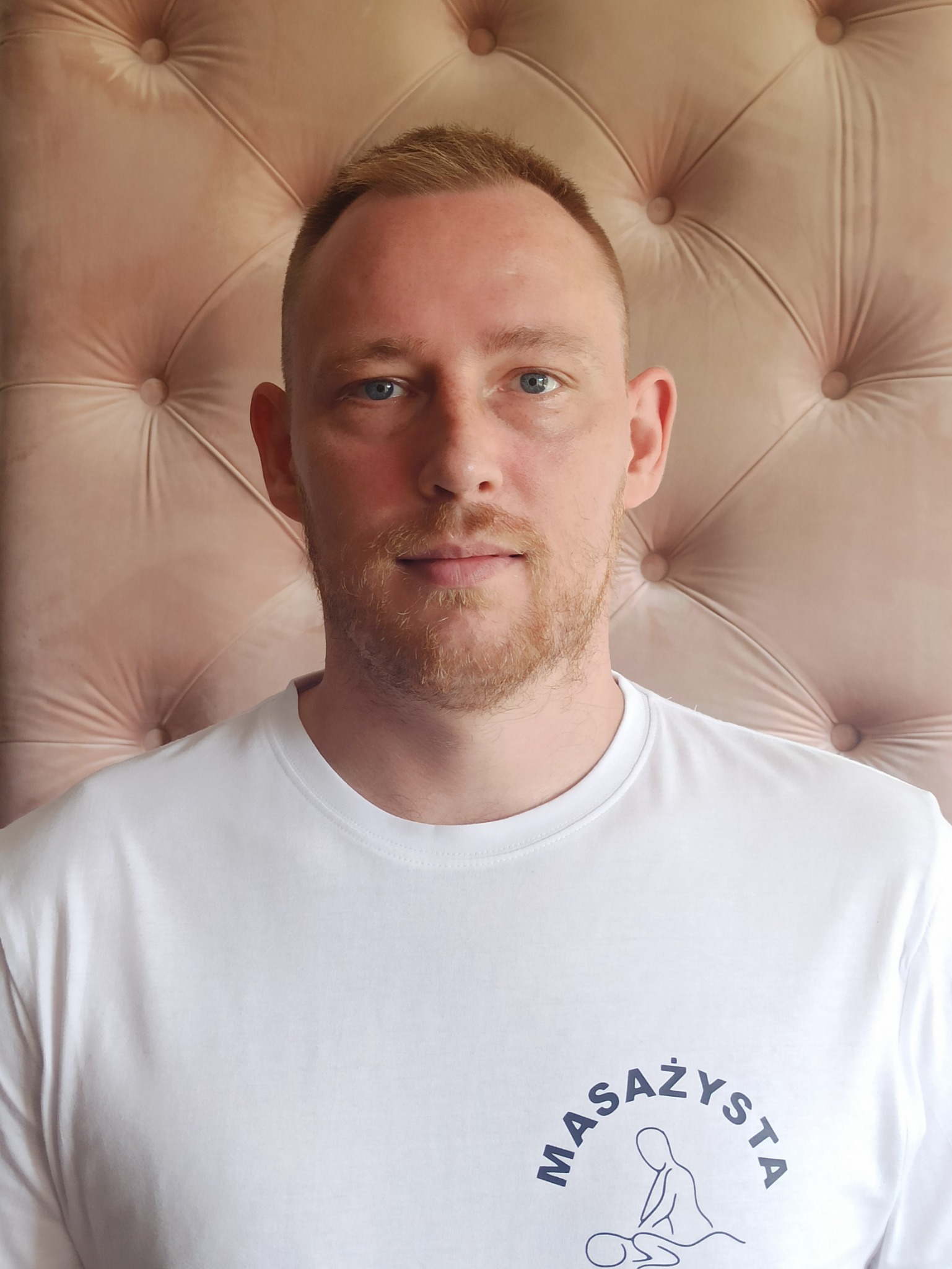 Portret mężczyzny w białej koszulce z logo firmy oferującej usługi masażu, na tle pikowanego, pudrowo-różowego zagłówka.