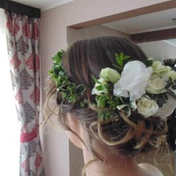 Wedding Assistance Kinga Rek - Upięcie ślubne z białych róż i zielonych liści, widok z tyłu, w tle okno z wzorzystymi zasłonami i fragment jadalni